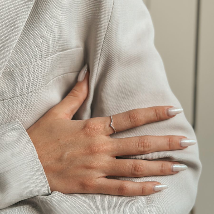 Közelkép egy kézről, amelyen Simple V ezüst gyűrű van, semleges színű textil háttérrel.
