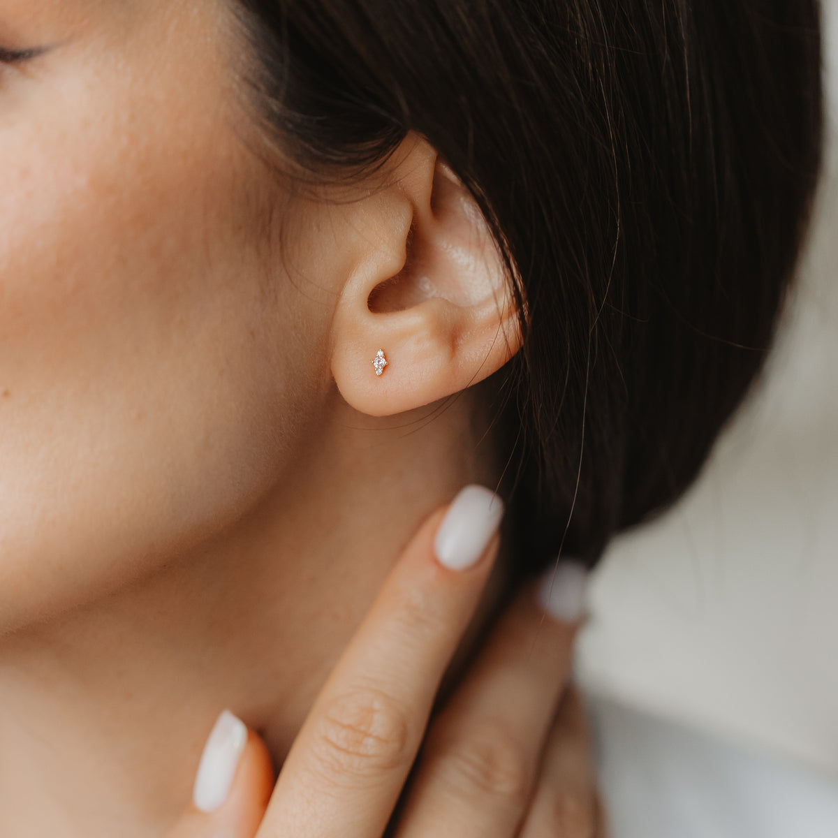 Közelkép egy fülről, amely apró, 14K sárga arany Flora bedugós fülbevalót visel rózsaszín morganittal és fehér zafírral, miközben egy semleges manikűrű kéz a nyakat érinti.
