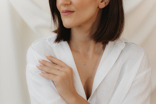 Közelkép egy fehér blúzt viselő személyről, aki finom 14K sárga arany gyűrűt visel rózsaszín turmalinnal és fehér zafírral; keze a vállán pihen, lágy, semleges háttér előtt.